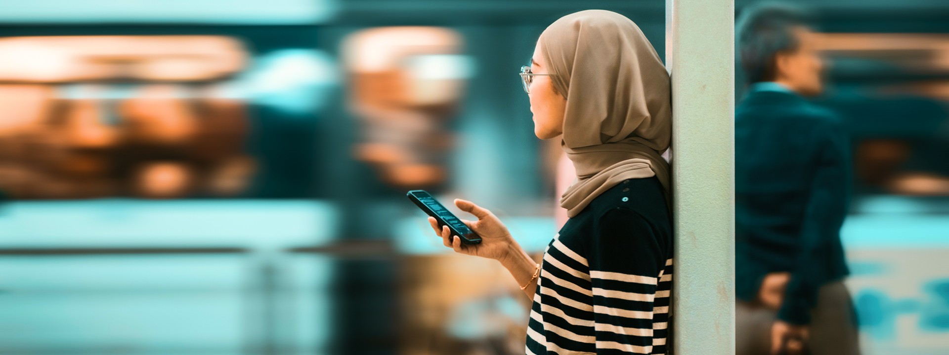 Woman in train station leans against wall while holding mobile phone