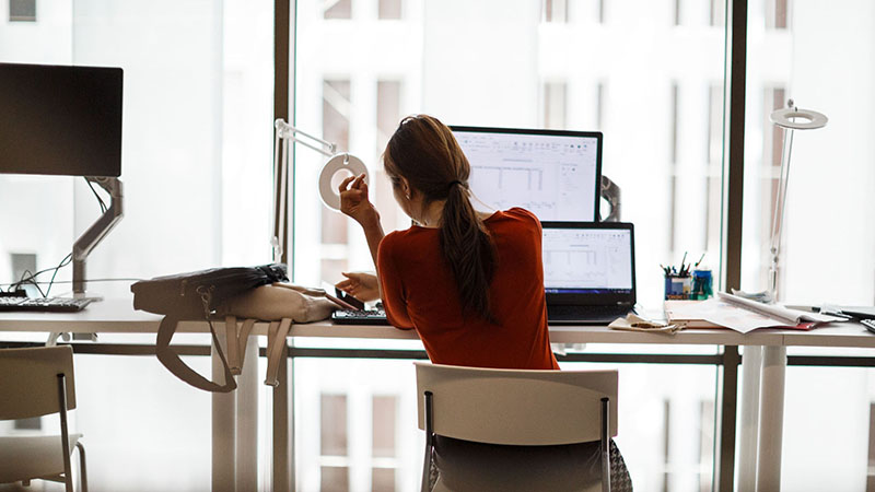 A woman sitting at a desk in front of a laptop and monitor with high-rise view.