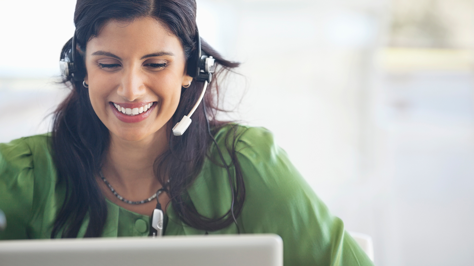 Mulher usando vestido verde, trabalhando em um laptop e utilizando headset de atendimento