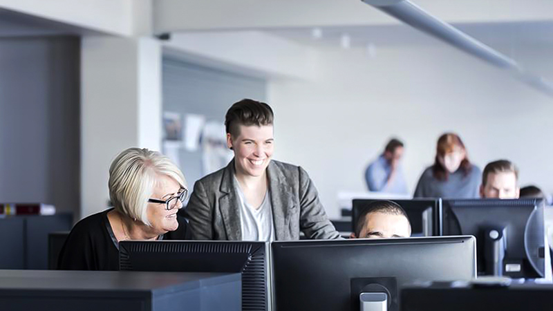 Colegas de trabalho felizes reunidos ao redor da mesa de um colega, discutindo o conteúdo exibido em seu computador