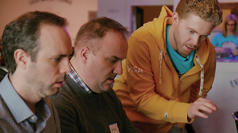 Three men, one in a Visa yellow jacket, another a gray sweater, and another in plaid, talk amongst each other. 