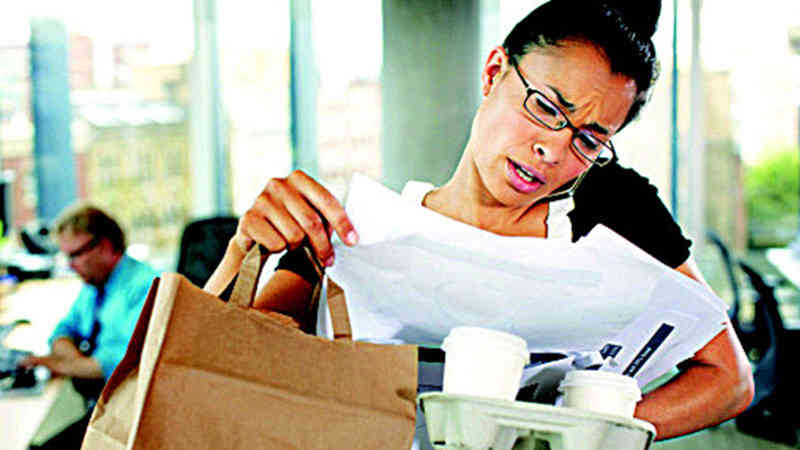 A businesswoman is speaking on a phone being held between her head and left shoulder with her right hand holding a paper bag and sifting through a pile of papers and her left hand holding a tray of to-go coffee cups.